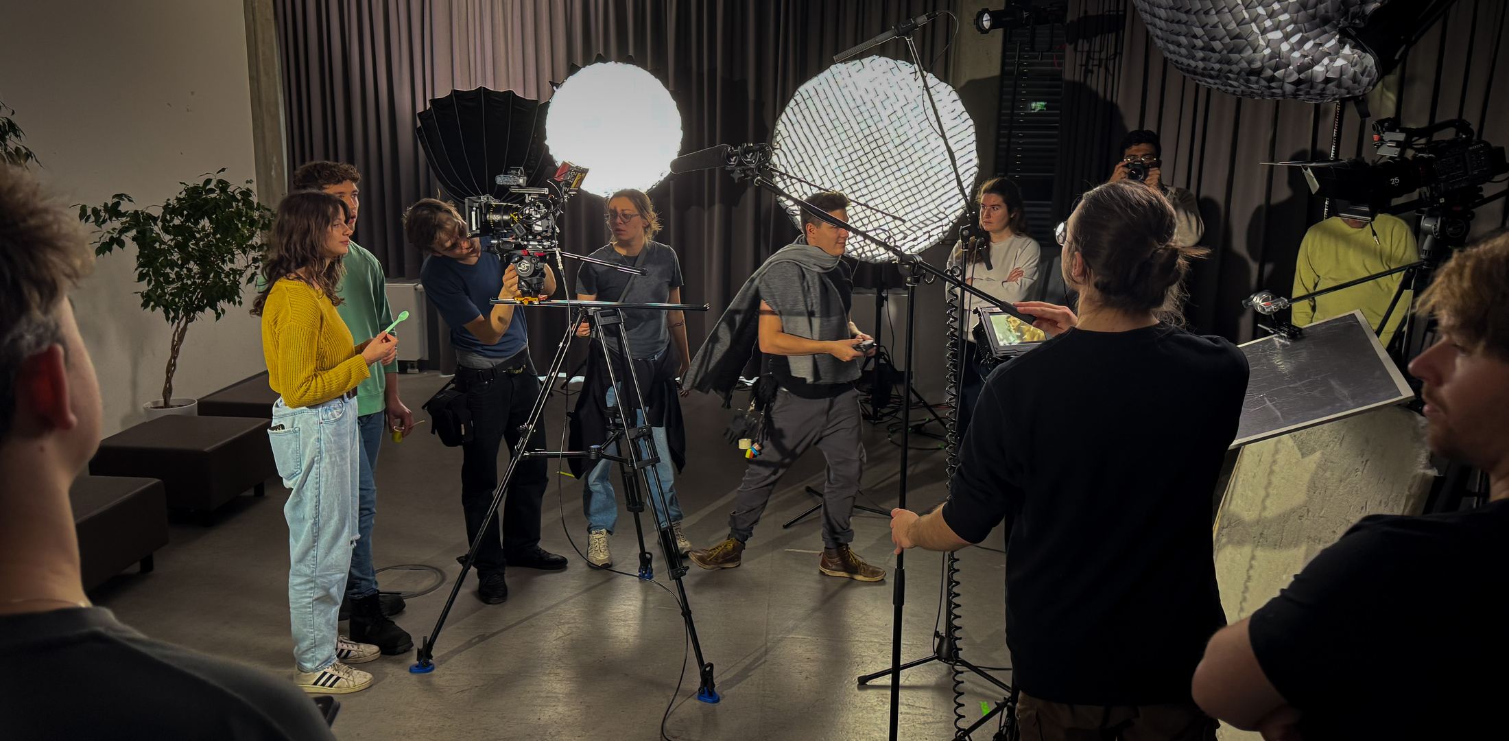 A Movie Sprint crew at work on set: two performers waiting, a camera operator on sticks, a boom operator, lights on stands and the rest of the crew watching from the edge of frame